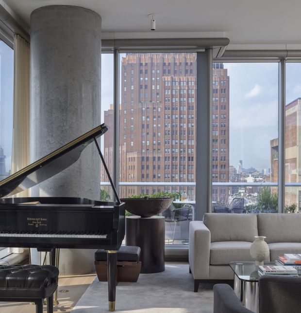 A sophisticated high-rise living room with floor-to-ceiling windows overlooking an urban skyline. The room features a black grand piano, a light upholstered sofa, dark lounge seating, a glass coffee table, sculptural planters, and a large exposed concrete column. The space has a refined contemporary aesthetic with abundant natural light.