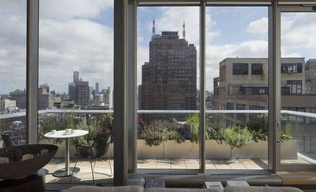 A bright contemporary apartment interior viewed through full-height windows, with a furnished balcony terrace featuring a small round table, chairs, and planter boxes. The glazing overlooks a dense urban skyline and a prominent historic high-rise building. A sofa is partially visible in the foreground.