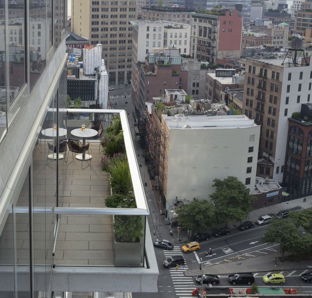 Elevated view over a dense urban city centre, with a narrow rooftop terrace in the foreground. The terrace has large-format grey paving, glass balustrades, raised planters with lush greenery, and a small round outdoor dining table with chairs. Surrounding mid- and high-rise commercial buildings, streets, crosswalks, and traffic are visible below.