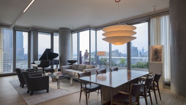 A sophisticated open-plan high-rise apartment interior combining a living room and dining area. Floor-to-ceiling corner glazing provides expansive urban views and access to a balcony. The space features a grand piano, sculptural layered pendant light, contemporary seating, a large timber dining table, dark wood dining chairs, polished concrete columns, and a restrained neutral palette.