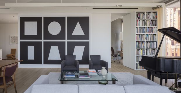 Bright, contemporary open-plan living room with a pale sectional sofa, two dark armchairs, glass coffee table, large geometric black-and-white wall art, floor-to-ceiling built-in book shelving, and a black grand piano. Timber flooring, white walls, and expansive glazing create an airy urban interior.