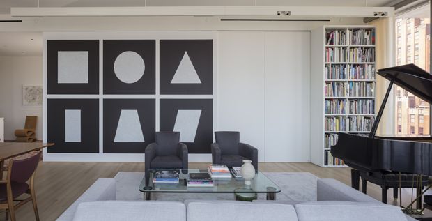 A bright, contemporary living room with a large pale sectional sofa, two dark upholstered armchairs, a glass coffee table, and a black grand piano. The room features light timber flooring, white walls and built-in cabinetry, a full-height bookcase filled with books, and a striking six-panel geometric artwork installation in black and grey. Large glazing at right provides an urban outlook, while a linear ceiling detail and recessed lighting contribute to the clean architectural character.
