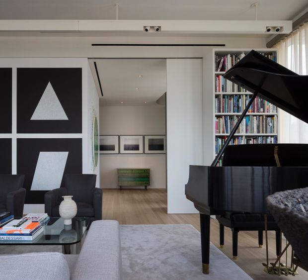 A refined contemporary living room featuring a glossy black grand piano, pale upholstered seating, a glass coffee table, large black-and-white geometric wall artwork, and a full-height bookshelf. The room opens toward a secondary gallery-like space with framed artwork and a low timber console. White walls, recessed linear lighting, pale timber flooring, and tall glazing create a bright, clean interior with strong black accents.