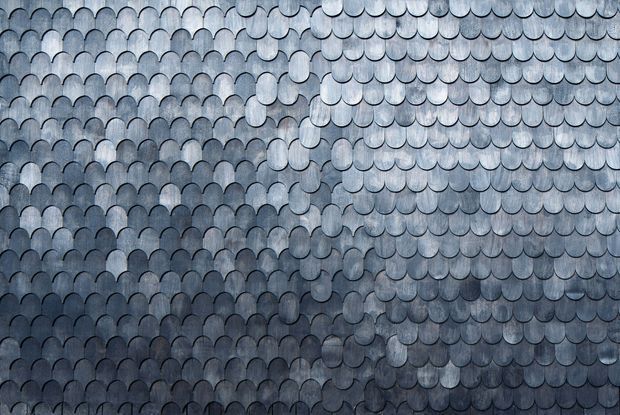 Close-up of an overlapping scalloped shingle or tile surface, likely a roof or cladding finish. The repeated rounded pieces create a fish-scale pattern, with varied blue-grey tones, dark seams, and a textured, weathered appearance. No room or broader building context is visible.