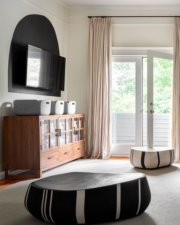 A bright, refined living room with a wall-mounted television set against a large arched black feature panel. A mid-century-inspired timber media cabinet with glazed doors and drawers sits below, accompanied by compact grey speakers. Full-height beige curtains frame an open glazed door to an exterior balcony or terrace. Two low, rounded black-and-white striped ottoman tables sit on a light carpet, creating a bold graphic accent.