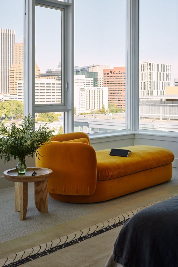 A bright, contemporary living room in a high-rise urban setting, featuring a sculptural mustard-orange velvet chaise lounge positioned beside large white-framed corner windows. A small timber side table with a glass vase and greenery sits nearby, while a patterned area rug and dark upholstered furniture add texture in the foreground. The windows overlook a dense city skyline.