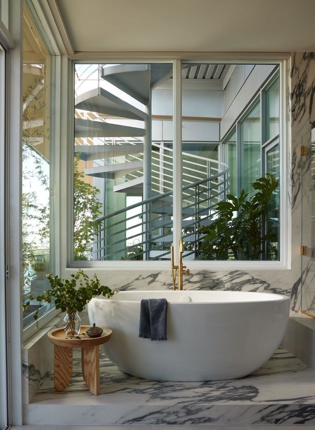 A bright, luxurious bathroom centered on a freestanding white bathtub positioned beside large floor-to-ceiling windows. The room features dramatic white marble with grey veining on the walls and floor, a brass floor-mounted bath filler, white-framed glazing, and a small timber side table with a plant. Through the window, a sculptural modern spiral staircase and adjacent glazed building elements are visible.