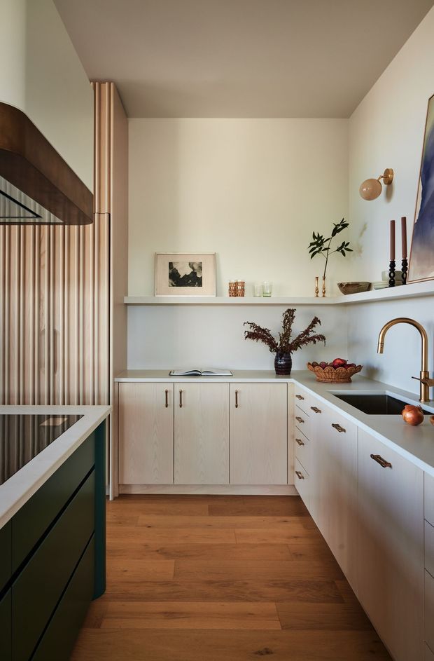 A warm, compact contemporary kitchen with pale timber cabinetry, white benchtops and splashback, and a central dark green island. The room features a timber floor, fluted timber wall screening, brass-toned hardware and tapware, open display shelving, decorative objects, and a framed artwork. Natural wood tones and muted finishes create a refined, inviting atmosphere.