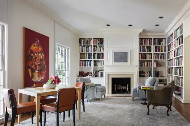 A bright, refined open-plan living and dining room with extensive built-in white bookshelves, a central white fireplace mantel, timber flooring, and large windows. A rectangular dining table with leather chairs occupies the foreground, while upholstered armchairs and a small seating arrangement sit around the fireplace. The room includes classic wall mouldings, recessed ceiling lights, artwork, and a large neutral rug.