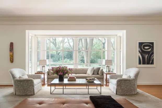 Bright, traditionally detailed living room with a large multi-pane bay-style window overlooking greenery. The room contains two upholstered armchairs, a sofa, a glass-topped rectangular coffee table, side tables with lamps, framed artwork, a leather ottoman, and a pale area rug. Warm timber flooring, neutral walls, substantial ceiling and window trim, and abundant natural light create a refined, inviting atmosphere.