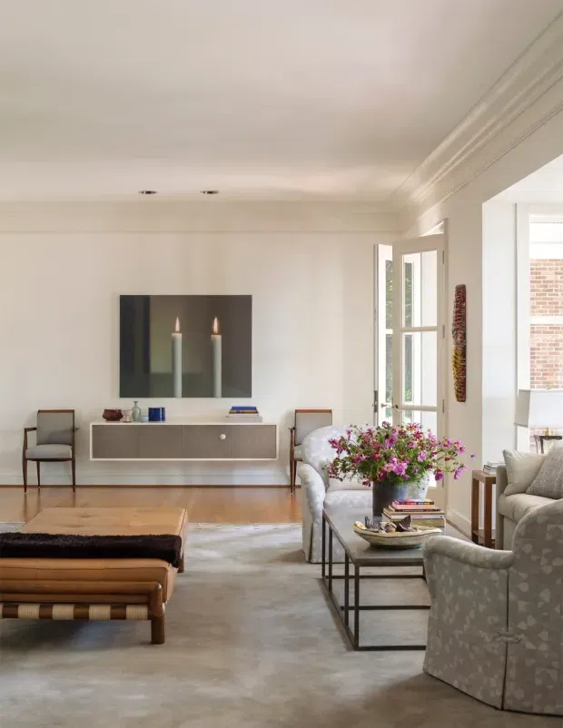 A refined, light-filled living room with white walls and detailed crown moulding, pale timber flooring, and a neutral upholstered seating arrangement. A low built-in floating console sits beneath a large artwork, while a central coffee table, patterned armchairs, fresh flowers, and layered books create an elegant, inviting setting. Glazed doors open toward a bright adjoining area.