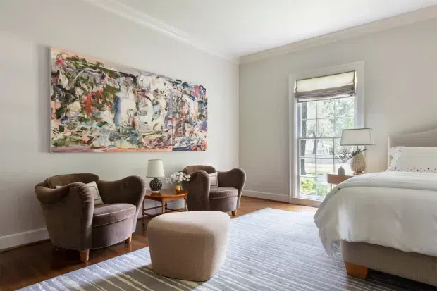 A bright, refined bedroom with a large upholstered bed, two plush armchairs, a round ottoman, and a small side table. The room has pale walls with decorative crown moulding, warm timber flooring, a large abstract artwork, a neutral area rug, and tall divided-pane glazing with a Roman blind.