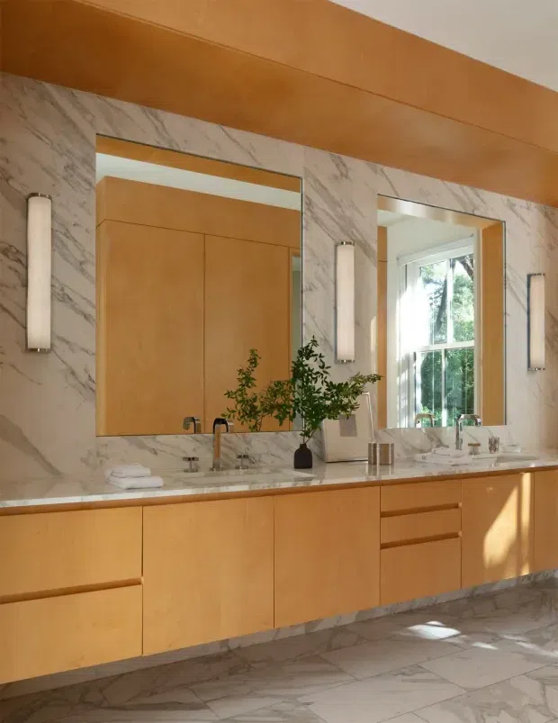 A refined bathroom vanity wall with two large mirrors, a long pale timber floating cabinet, twin basins and polished metal tapware. Dramatic veined marble covers the wall and floor, while slim vertical wall sconces and warm timber detailing create a clean, luxurious atmosphere. Natural light enters through a window reflected in the right mirror.
