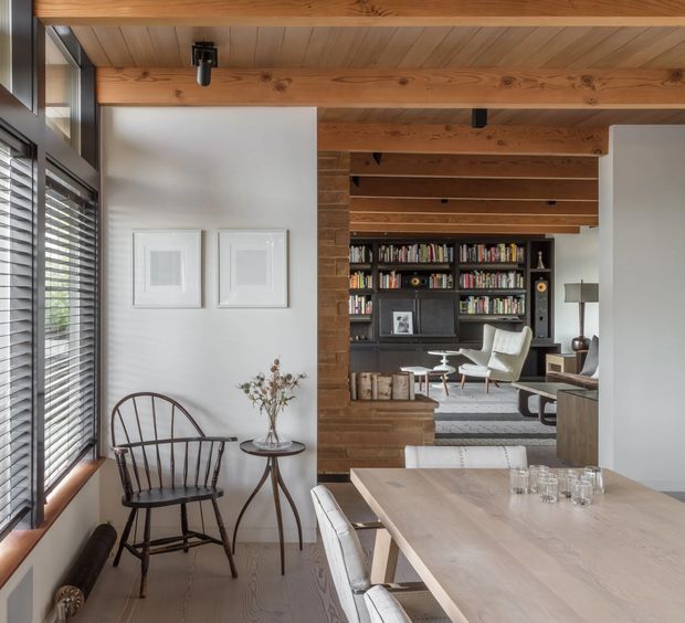 A warm, wood-lined open-plan interior combining a dining area with a living room beyond. The foreground features a light timber dining table and upholstered chairs beside a large window with horizontal blinds, while exposed timber ceiling beams, timber flooring, and a dark built-in bookcase filled with books define the layered living space. A spindle-back armchair, side tables, framed artwork, and a lounge seating area create a refined, inviting atmosphere.