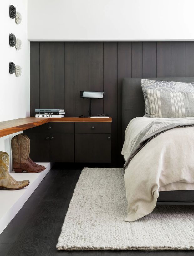 A refined bedroom interior featuring a bed with layered neutral grey and cream bedding, a dark vertical-panel feature wall, and integrated dark timber cabinetry beside the bed. A warm timber desktop or console extends from the cabinetry, topped with books and a small lamp. A white built-in display ledge holds decorative boots, while a textured white rug covers part of the dark timber floor. The overall design is contemporary with restrained, neutral styling and strong contrast between dark joinery and light surfaces.