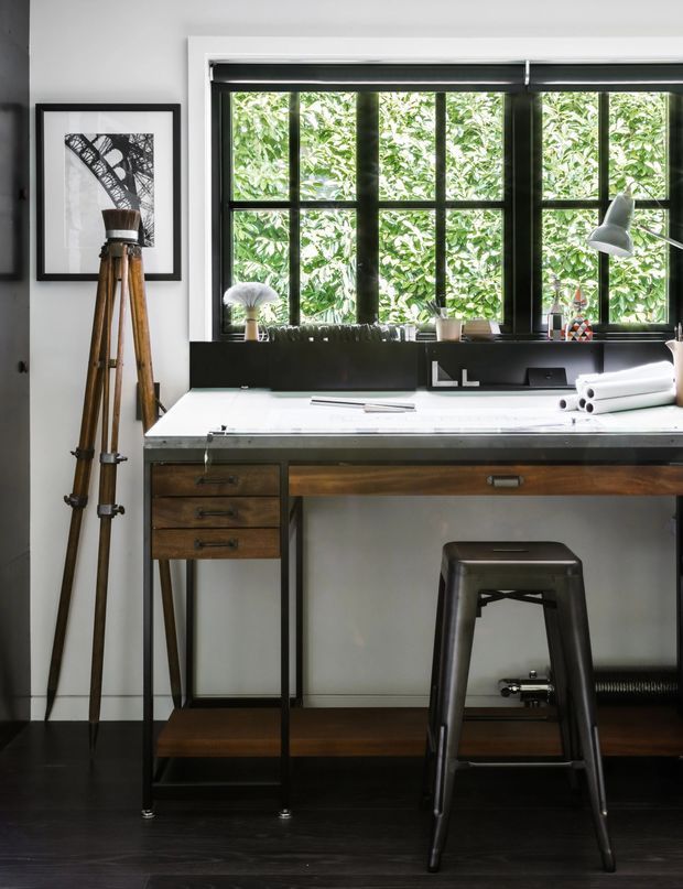 A stylish home office or drafting workspace with a long built-in-style timber and black metal desk, white work surface, drawers, shelving, and a dark stool. A large black-framed multi-pane window overlooks dense greenery, while a desk lamp, rolled drawings, drafting tools, and framed artwork reinforce the creative studio character. The room has dark flooring, white walls, and a restrained industrial-contemporary palette.