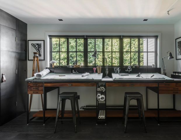 A stylish compact home office with a long built-in workbench positioned beneath large black-framed multi-pane windows. The room features dark timber flooring, charcoal cabinetry and shelving, a pale worktop, two industrial-style stools, drafting materials, task lamps, and neatly arranged storage. Lush greenery is visible outside the windows, creating a calm, light-filled workspace.