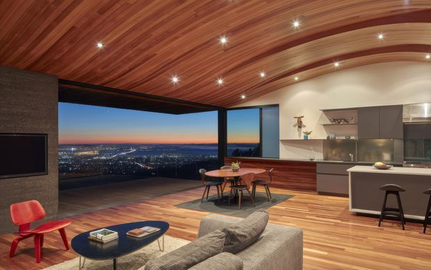 An open-plan contemporary living, dining, and kitchen interior with expansive glazed corner views over a city at dusk. The room features a dramatic curved timber-lined ceiling with recessed spotlights, timber flooring, a dark stone or textured feature wall with integrated television, a sectional sofa and coffee table, a round dining table, and a streamlined kitchen with dark cabinetry and a central island.