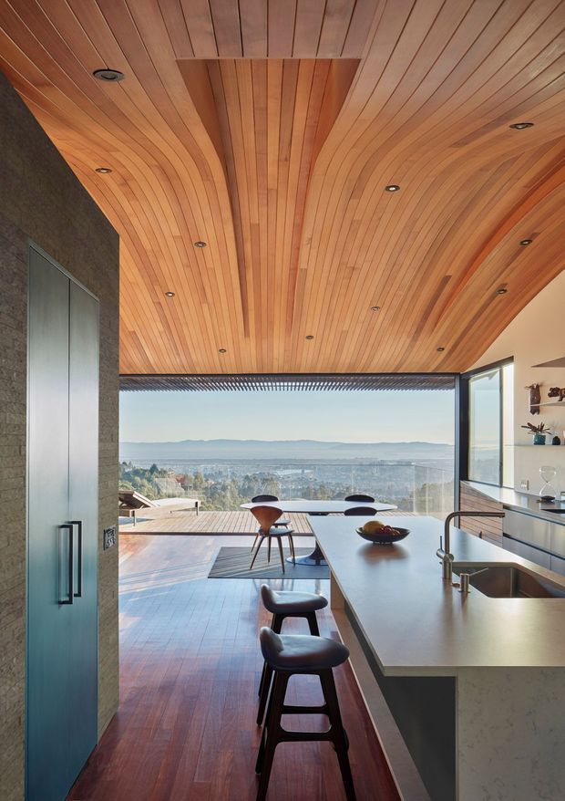 A contemporary open-plan kitchen and living area with a dramatic curved timber-lined ceiling and warm timber flooring. A large stone island with an integrated stainless-steel sink and dark stools runs toward full-height glazing, opening to a timber deck and expansive distant water and landscape views. Dark textured wall cabinetry, recessed downlights, and refined minimal furnishings create a warm, architectural interior.
