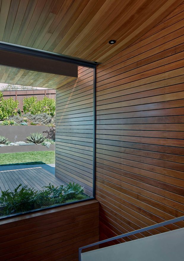 A contemporary timber-lined stairway or landing with warm horizontal wall panelling and a matching slatted ceiling. A large corner window overlooks a landscaped courtyard with raised garden beds and succulents, while dark metal window framing, recessed lighting, and a minimalist stair balustrade add contrast.