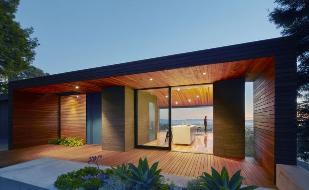 Contemporary single-storey pavilion viewed from an outdoor deck, with dark horizontal exterior cladding framing a recessed timber-lined entry and large floor-to-ceiling glazed opening. Warm timber decking and ceiling, dark structural walls, landscaped planting, and a bright interior with a kitchen island are visible; a waterfront or ocean view appears beyond the glazing.