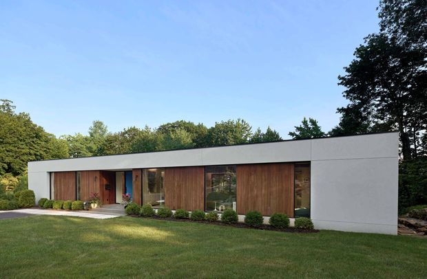 A low, single-storey modern house set within a landscaped garden. The long rectangular facade combines smooth white rendered wall sections with warm vertical timber cladding, recessed glazing, and a flat roofline. Neatly clipped shrubs line the front elevation, with a broad lawn and mature trees surrounding the building.