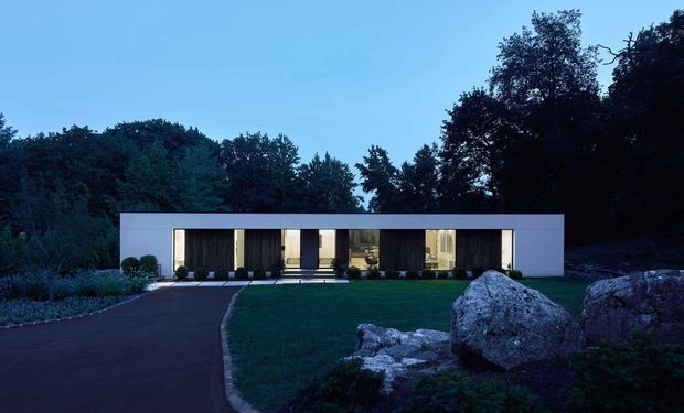 A low, rectilinear modern house set within a wooded landscape at dusk. The minimalist white facade features a broad flat roofline, tall recessed glazed openings, dark vertical wall panels, and a long sheltered entrance or living frontage. A curved paved driveway and large natural boulders occupy the foreground, with dense mature trees surrounding the property.