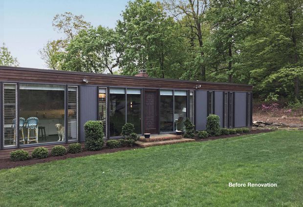 Single-storey, low-profile rectangular dwelling set in a wooded residential landscape, photographed from the lawn before renovation. The facade has dark vertical cladding, extensive floor-to-ceiling glazing with dark frames, a shallow flat or very low-pitched roof, and a small brick entry step. Neatly clipped shrubs and lawn occupy the foreground; indoor dining furniture and a dog are visible through the windows.