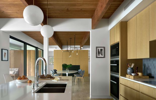 A bright contemporary open-plan kitchen viewed from the island, with pale timber cabinetry, a white stone island benchtop, integrated black appliances, and a grey tiled splashback. The space opens to a dining and living area with large black-framed glazing. Exposed timber ceiling boards and substantial timber beams add warmth, while spherical pendant lights and slim black dining pendants provide modern detailing.