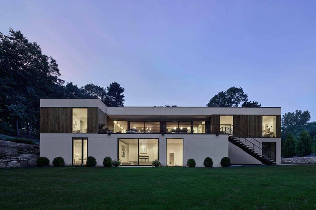 A dusk exterior view of a large, modern two-storey residence set within a landscaped, tree-lined site. The building has a strongly horizontal composition with flat roofs, white rendered walls, dark timber-clad sections, extensive glazing, an upper-level terrace or walkway, and a prominent external staircase. Warm interior lighting contrasts with the cool blue evening sky.