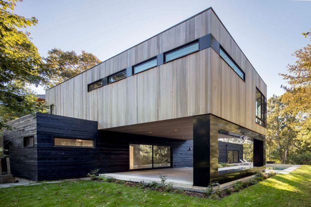 A contemporary two-storey residential house with a dramatic cantilevered upper level. The facade combines vertically clad light timber with dark horizontal cladding at the lower level, large horizontal strip windows, and substantial dark structural columns supporting the overhang. The home is set within a leafy landscaped suburban garden, with a partially covered outdoor terrace and glazing opening toward the yard.