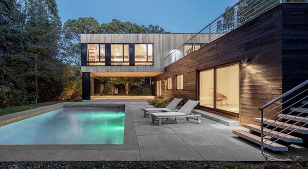 Contemporary two-storey residence viewed from a landscaped pool terrace at dusk. The home combines dark horizontal timber cladding with a pale upper volume, extensive glazing, a covered outdoor passage, rooftop or upper-level metal balustrading, and an angular exterior stair. A rectangular illuminated swimming pool is surrounded by large grey paving, with outdoor loungers beside the timber-clad facade.