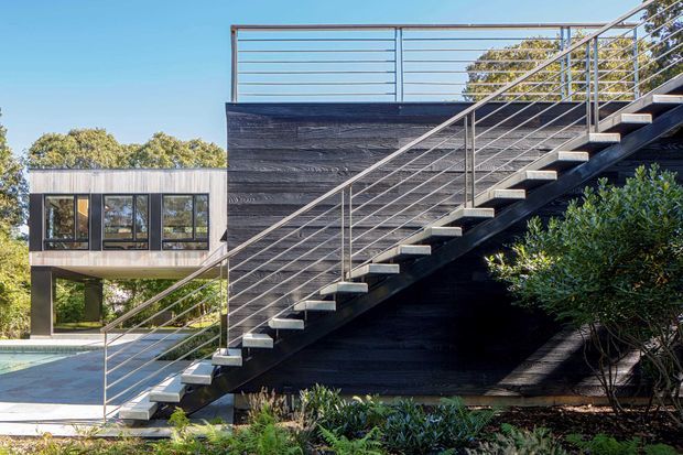 A contemporary multi-level building exterior with a prominent open-riser metal staircase and slim horizontal railings climbing alongside a dark textured wall. A glazed upper-level volume, flat rooflines, planted landscaping, and part of a swimming pool are visible in bright daylight.