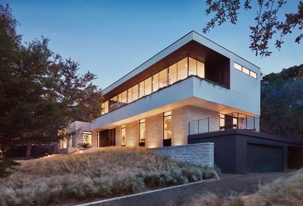 Twilight exterior view of a contemporary multi-level residence set among mature trees and landscaped grasses. The home has strong horizontal volumes, a low-pitched asymmetrical roof, extensive floor-to-ceiling glazing, warm timber soffits, pale rendered and masonry walls, dark metal framing, and an integrated double garage. A balcony or terrace projects from the upper level, with warm interior lighting visible through the glazing.