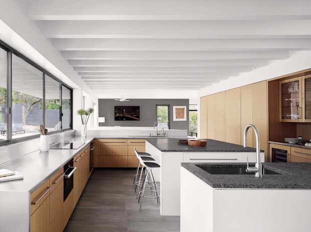 Bright contemporary open-plan kitchen with extensive warm timber cabinetry, dark stone benchtops, a white island with integrated sink, and white bar stools. The room features large black-framed glazing, a white exposed-beam ceiling, and an adjoining living area visible beyond the kitchen.