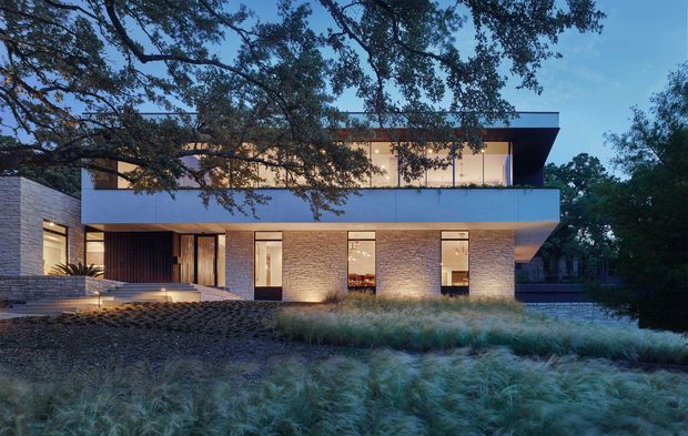 Twilight exterior view of a contemporary two-storey residence set within landscaped ornamental grasses and mature trees. The building has a strong horizontal flat-roofed form, projecting upper level, extensive glazed openings, warm brick masonry walls, dark timber or metal screening at the entry, and subtle architectural lighting.