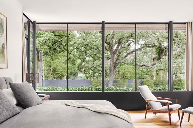 A bright, refined bedroom with a large upholstered bed in the foreground and a mid-century-style lounge chair beside it. Floor-to-ceiling black-framed glazing spans the rear wall, overlooking leafy trees and a landscaped garden. The room has pale walls, light timber flooring, neutral textiles, dark window framing, and understated contemporary furnishings.