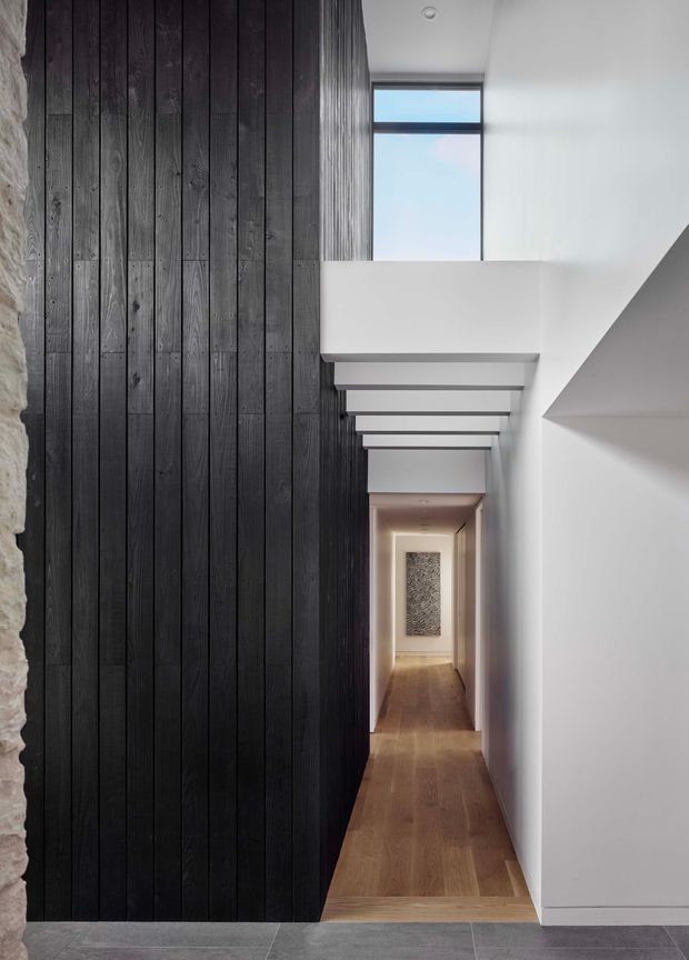 A narrow, dramatic interior hallway with a long dark vertical timber-panelled wall on the left, smooth white walls and a white angular ceiling on the right, and a series of exposed ceiling beams creating a rhythm toward the far end. Warm timber flooring leads to a softly lit passage with artwork, while a high narrow window brings in natural light. A rough stone edge is visible at the entrance and large grey floor tiles transition into the timber floor.