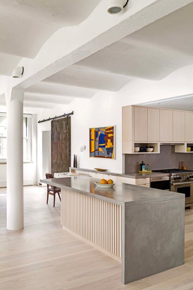Bright open-plan kitchen with a large central island, pale timber cabinetry, grey concrete-look benchtops, and a vertically fluted island front. The room features white plaster walls and a sculptural textured ceiling with exposed beam-like forms, pale timber flooring, integrated appliances, a freestanding stainless-steel range, a sliding barn-style door, and colourful abstract artwork. A dining area and tall windows are visible in the background.