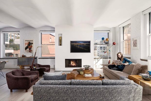 Bright, spacious contemporary living room in an urban apartment, featuring exposed white-painted brick walls, large industrial-style windows, a central fireplace with television above, and layered seating arranged around a round timber coffee table. The room includes a sofa, upholstered lounge chairs, a small desk area, and abundant natural light with city buildings visible outside.
