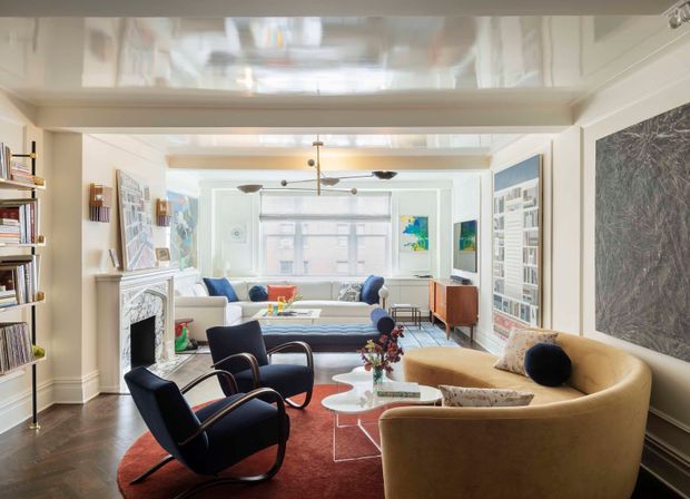 A bright, refined living room with a layered seating arrangement, including a large curved tan sofa, dark sculptural lounge chairs, and a pale blue sofa. The room features built-in bookcases, a decorative fireplace surround, artwork, a large red area rug, glass coffee tables, dark timber flooring, detailed wall panelling, and a glossy reflective ceiling with exposed beams and contemporary pendant and ceiling lighting.