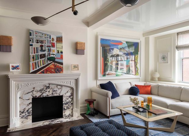 Bright, refined living room with a large white decorative fireplace surround featuring marble-patterned panels, a cream sectional sofa, tufted dark ottoman, glass-top geometric coffee table, colourful artwork, built-in shelving, and sculptural contemporary ceiling lighting. The room combines traditional detailing with eclectic modern furnishings and artwork.