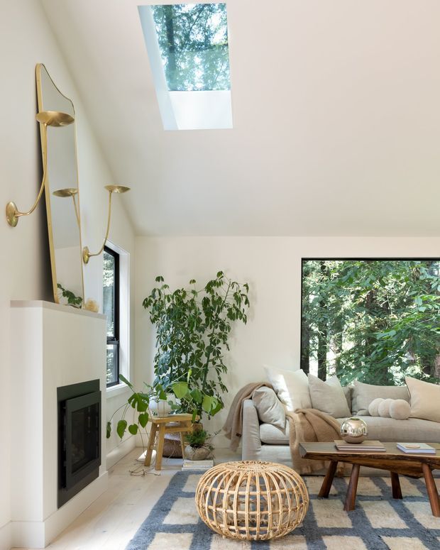 Bright, airy living room with a white fireplace surround and black inset firebox, a large beige sectional sofa, timber coffee table, woven rattan ottoman, patterned rug, indoor plants, and sculptural brass wall lighting. A skylight and large windows bring in natural light and views of trees. The room has a clean contemporary feel with warm natural accents.