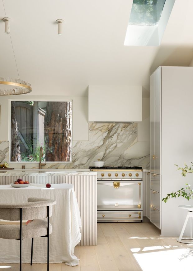 Bright, refined kitchen interior with a pale neutral palette. The room features white built-in cabinetry, a large veined marble splashback, a classic freestanding range cooker with brass controls, a fluted island, pale timber flooring, a small dining table with linen tablecloth, and upholstered stools. A skylight and large window provide abundant natural light, while a sculptural pendant and indoor plants add decorative detail.