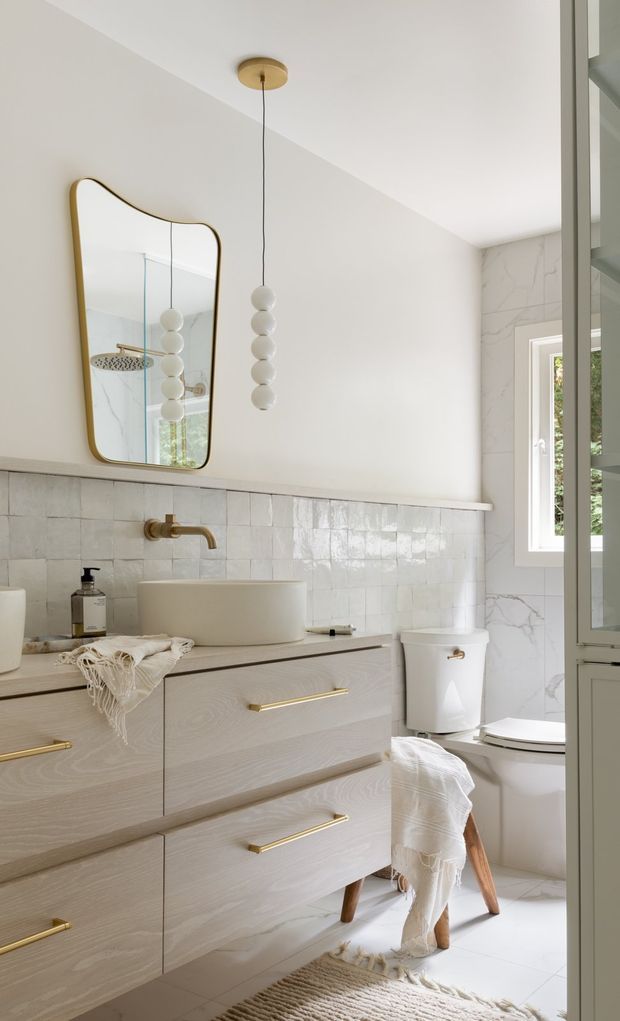 Bright, softly styled bathroom viewed from the doorway, featuring a pale timber floating vanity with long brass handles, white vessel basins, a brass wall-mounted tap, glossy square wall tiles, and large white marble-look tiles around the window and toilet. A sculptural gold-framed mirror and clustered globe pendant add decorative detail, while a partially visible shower appears in the mirror reflection. Natural light, warm neutral finishes, and woven textiles create an airy, refined atmosphere.