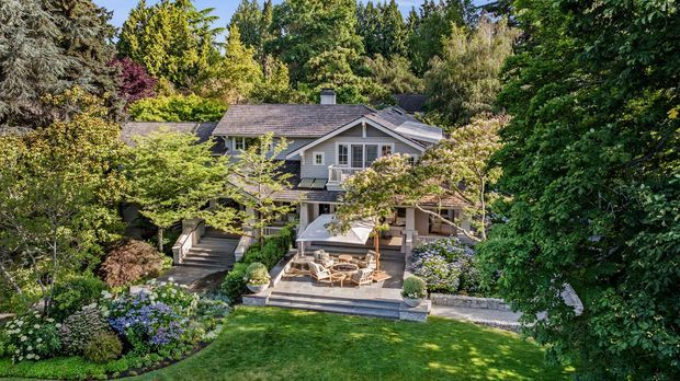 A landscaped residential house viewed from the garden, featuring a grey shingle-style pitched roof, light-coloured exterior walls, multi-level timber decks and steps, and a covered outdoor seating area. Mature trees, layered planting, flowering shrubs, stone garden edging, and a broad lawn surround the home.