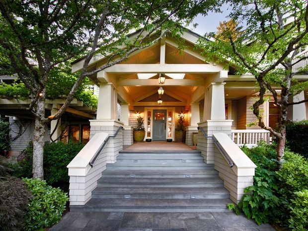 Front exterior view of a grand residential entry porch, approached by broad grey stone steps and flanked by white-painted masonry stair walls and columns. The covered entry has a warm timber-lined gabled ceiling, exposed beams, pendant lanterns, and glazed double doors. Mature trees and dense landscaping frame the facade, with glimpses of horizontal weatherboard cladding on the surrounding house.