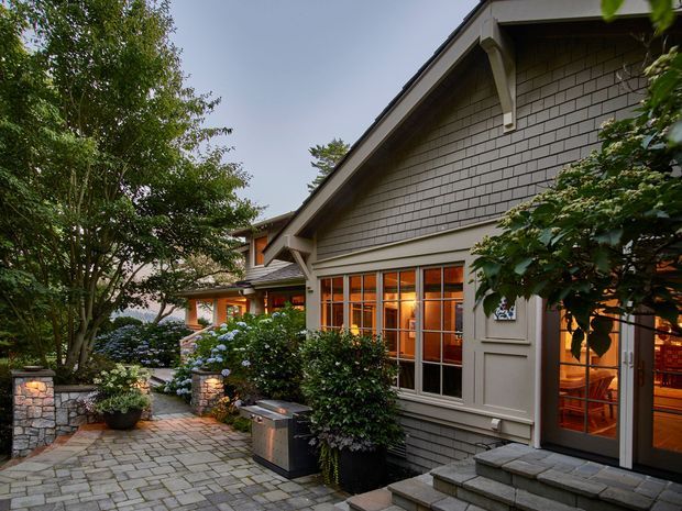 Exterior view of a residential home with a steep gabled roof, grey shingle-clad walls, white trim, large glazed doors and windows, and a landscaped stone-paved courtyard. Warm interior lighting is visible through the glazing, with mature trees, shrubs, hydrangeas, and low stone garden walls surrounding the entry.