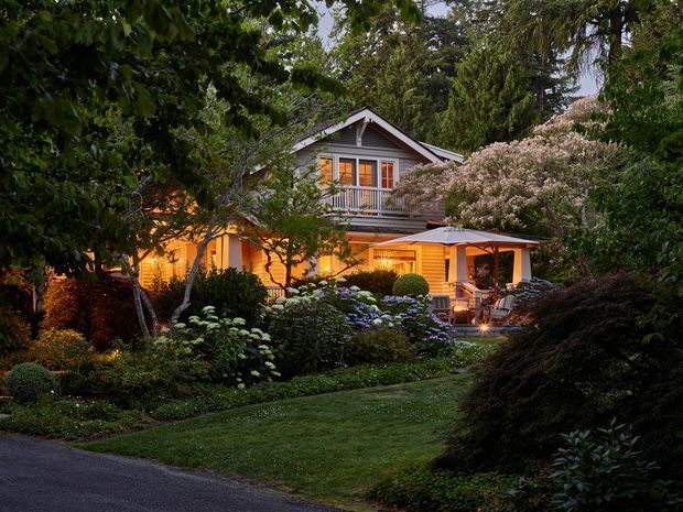 A warmly illuminated multi-level house nestled within a lush, mature garden. The home has a gabled upper roof, light timber or weatherboard cladding, large glazed doors and windows, and a covered outdoor terrace or patio with seating. Dense trees, shrubs, hydrangeas, manicured lawn, and garden lighting create an intimate evening setting.