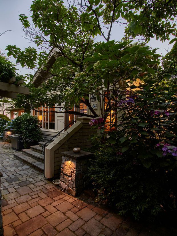 A warmly lit residential exterior viewed through dense mature garden planting. The scene shows a paved brick walkway and steps leading to a porch or entry, with low stone retaining walls, timber or painted architectural elements, glazed doors and windows, and illuminated stone pillars. Purple flowering shrubs and leafy trees frame the building, creating a sheltered, intimate approach.